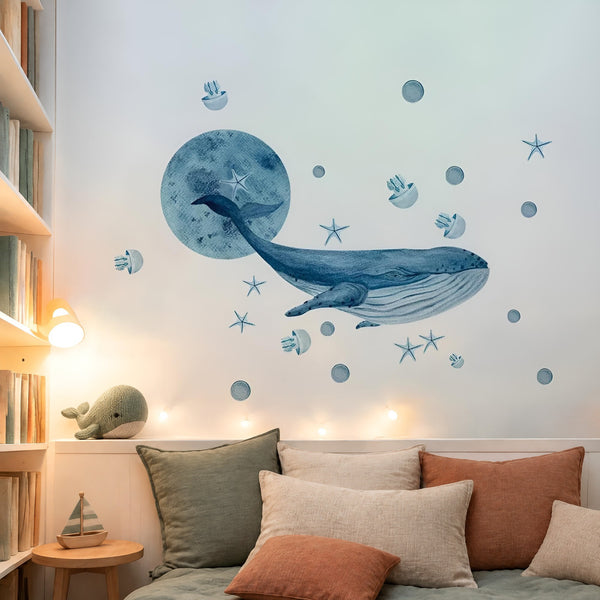 Grande adesivo murale balena ad acquerello e luna blu sopra una panca con cuscini in lino verde salvia, crema e ruggine in un angolo lettura