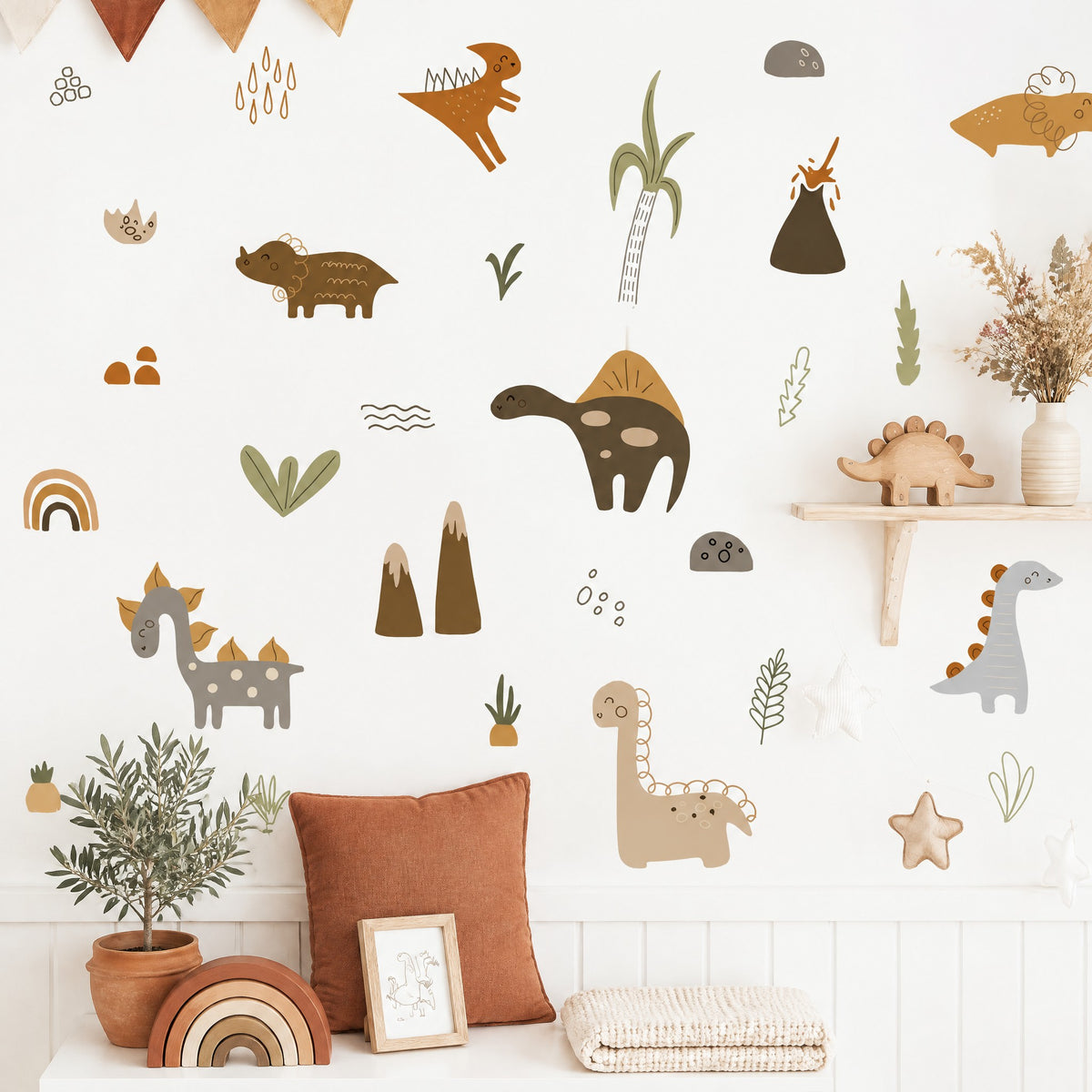 Stickers dinosaures, montagnes et feuillages aux tons de terre au mur, coussin rouille, olivier en pot de terre cuite et arc-en-ciel en bois