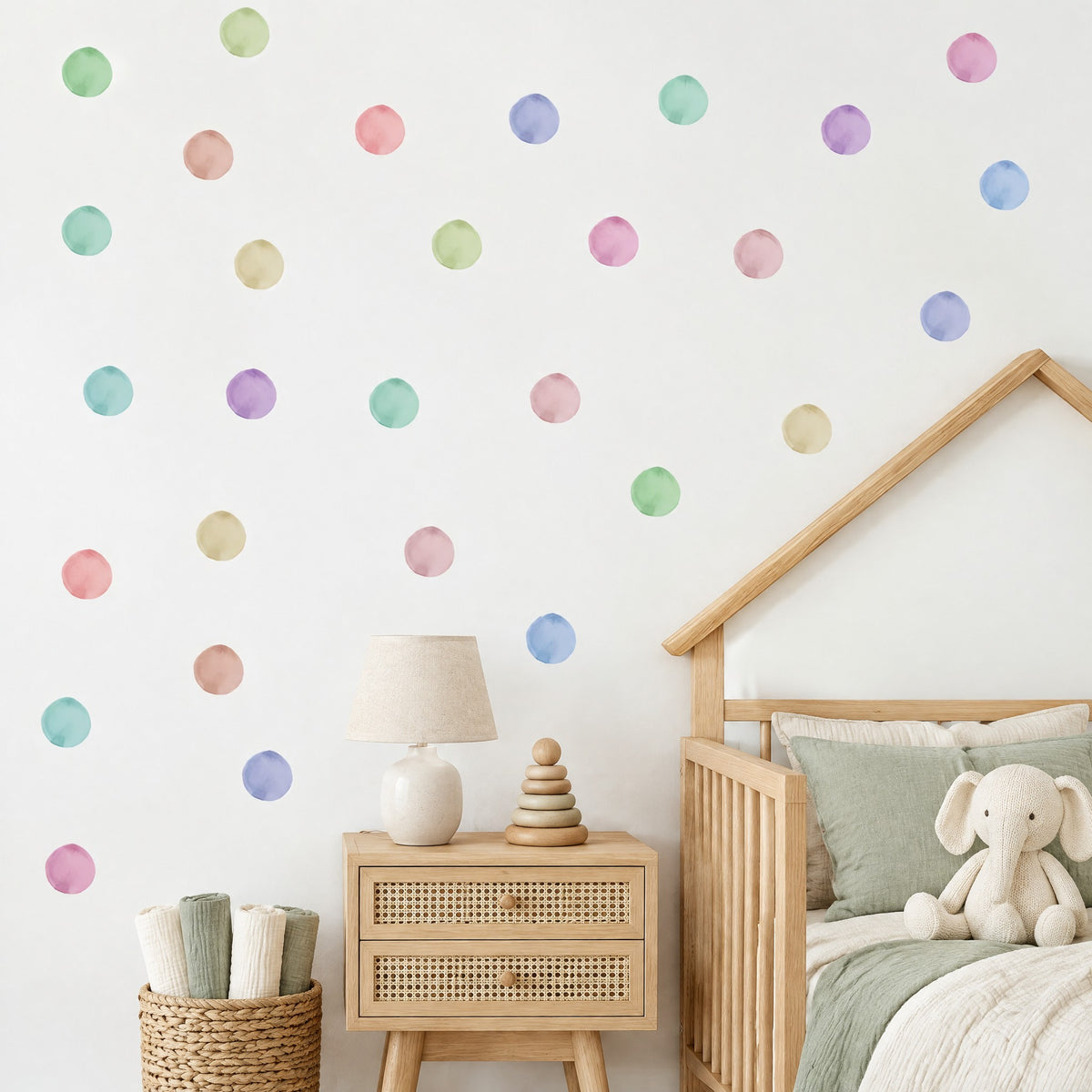Stickers muraux pois ronds aquarelle pastel sur un mur blanc de chambre de bébé, au-dessus d'un chevet canné et d'un lit cabane à barreaux en bois clair