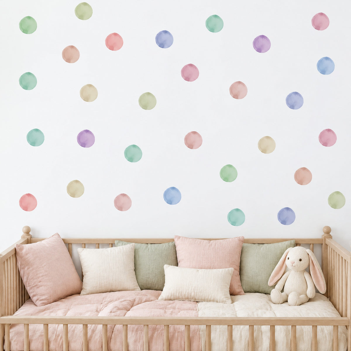 Pois aquarelle pastel au mur au-dessus d'un lit bébé à barreaux en chêne clair garni de coussins en lin rose poudré, crème et sauge