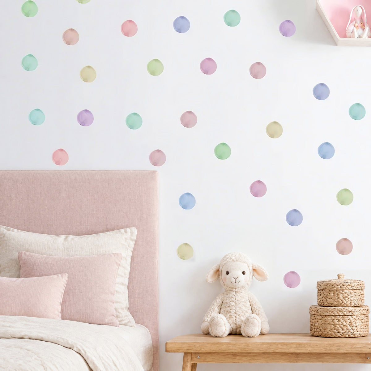 Pois aquarelle rose, vert, bleu et lavande sur le mur d'une chambre de bébé fille, au-dessus d'une tête de lit en lin rose poudré, d'un banc en chêne et d'un agneau en peluche